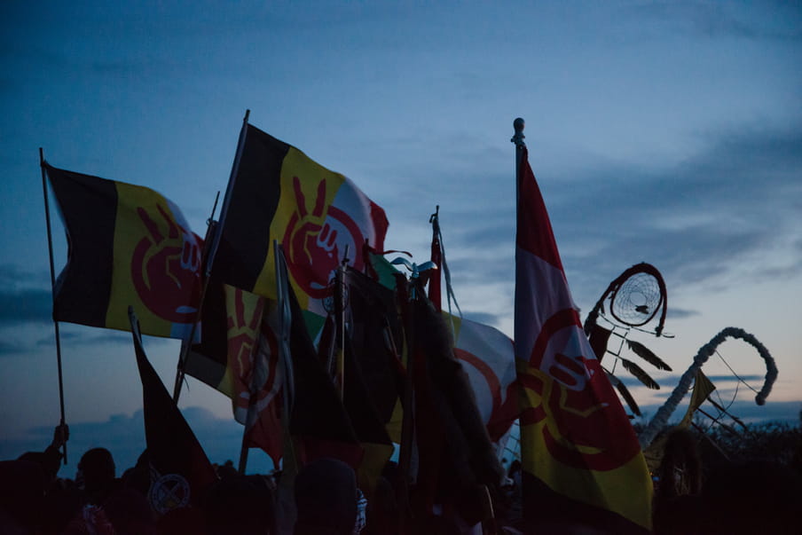 Photo of people holding up flags with a black, yellow, white and red stripe. In the middle theres a hand holding up two fingers.