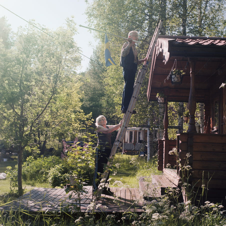 Photograph of an elderly couple on a ladder that stands against a wooden cabin. The man is holding a Swedish flag, that he want to attach to the cabin. The scene takes place in a green scenery with trees in the background and white flowers in the foreground. 