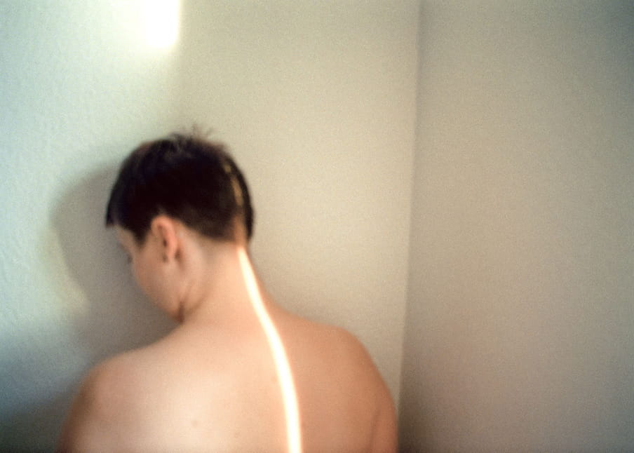 Slightly blurry photo of a person with short, black, spiky textured hair facing a wall in the corner of a room, we see the side of their face, with a long ray of sunlight running up their back and across their head 
