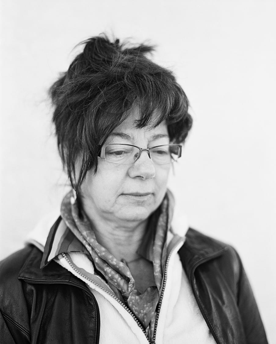 Black and white photo side profile of a person with short hair, falling around forehead and side of face as fringe, with glasses and a scarf collar, in a black leather jacket and zip up flece, eyes looking off to the right and down