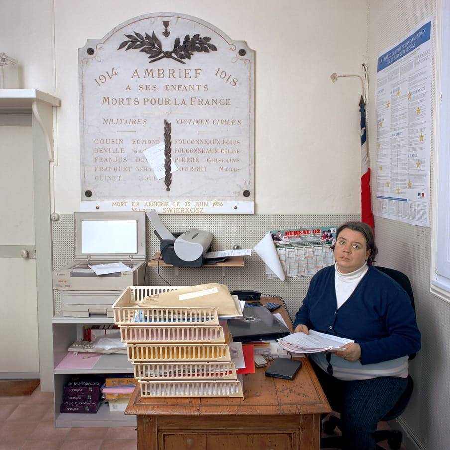 Female town clerk sits at a worn, wooden desk. She wears jeans and a navy blue, striped cardigan over a white, collared t-shirt. The desk has a yellow filing system filled with paper and envelopes. We also see a calculator and a closed laptop. There’s a copy machine and a fax machine at the back of the office. A pole with the French flag leans against the corner. A large, light-grey WWI memorial artwork made of marble, with engraved names of deceased soldiers and civil persons, is attached to the wall.