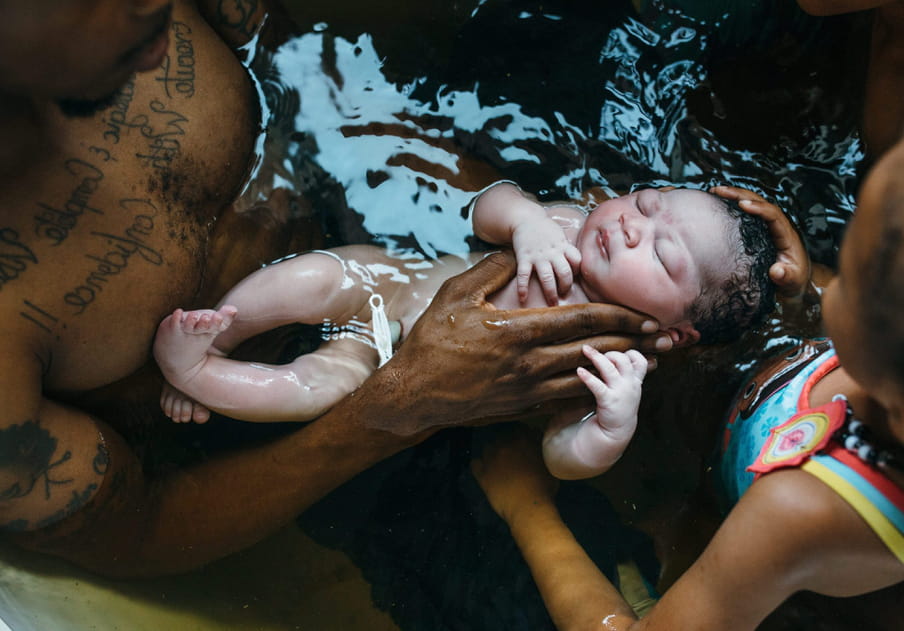 A newborn baby with a clamped umbilical chord is seen in water held by a man and a girl.
