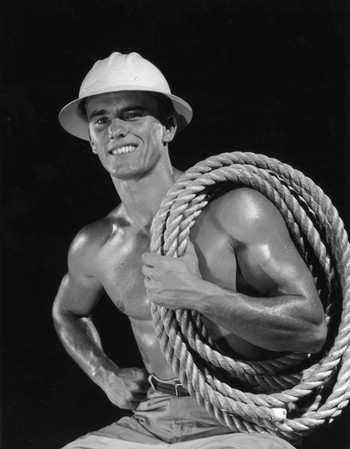Studio portrait of a bare-chested construction worker with glistening muscles, wearing a hard hat and holding thick rope coiled over one shoulder. Photo by Lambert / Getty Images